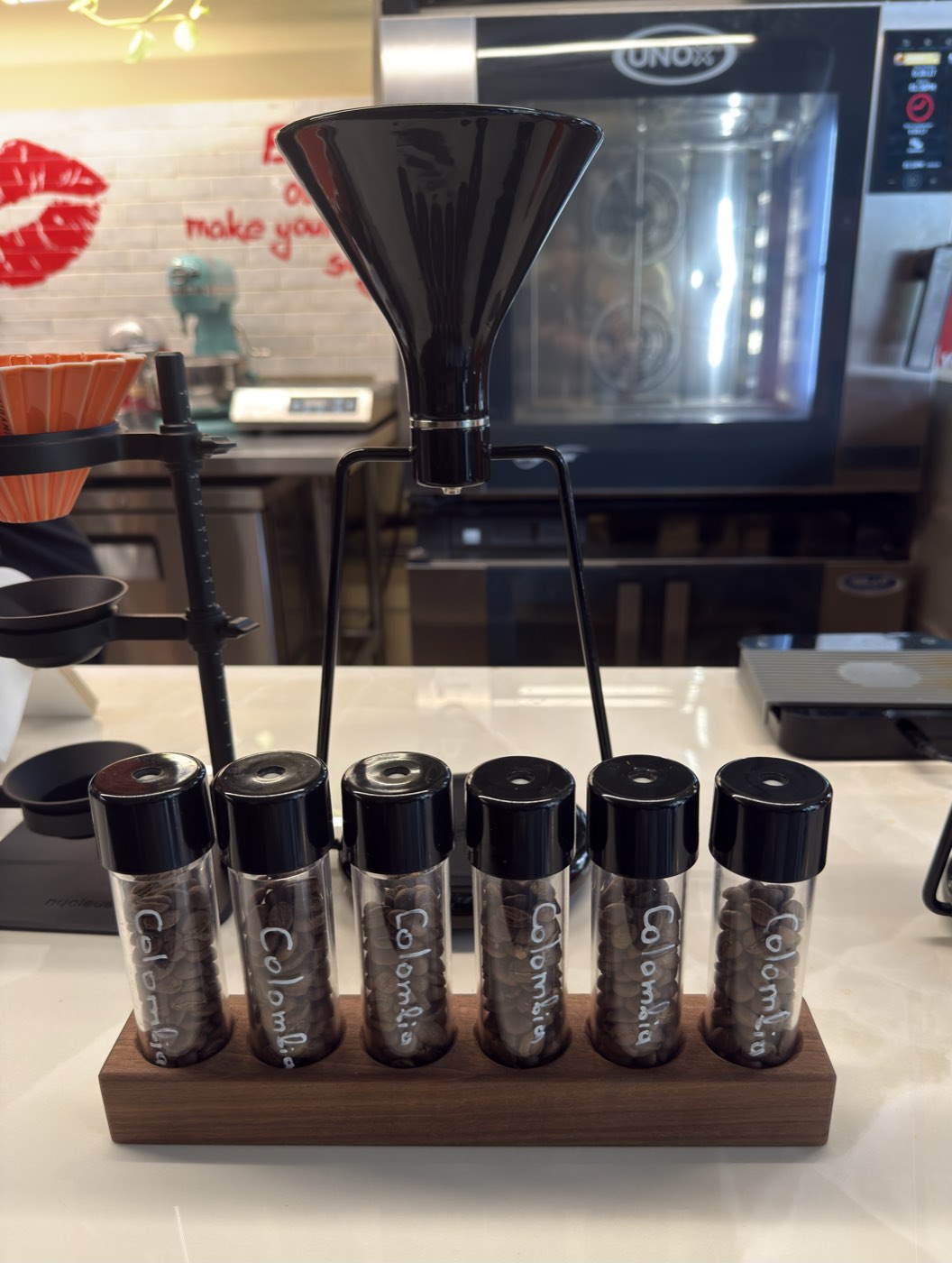 Six glass vials of single-origin coffee beans labelled Colombia on a wooden stand, with a black pour-over funnel behind