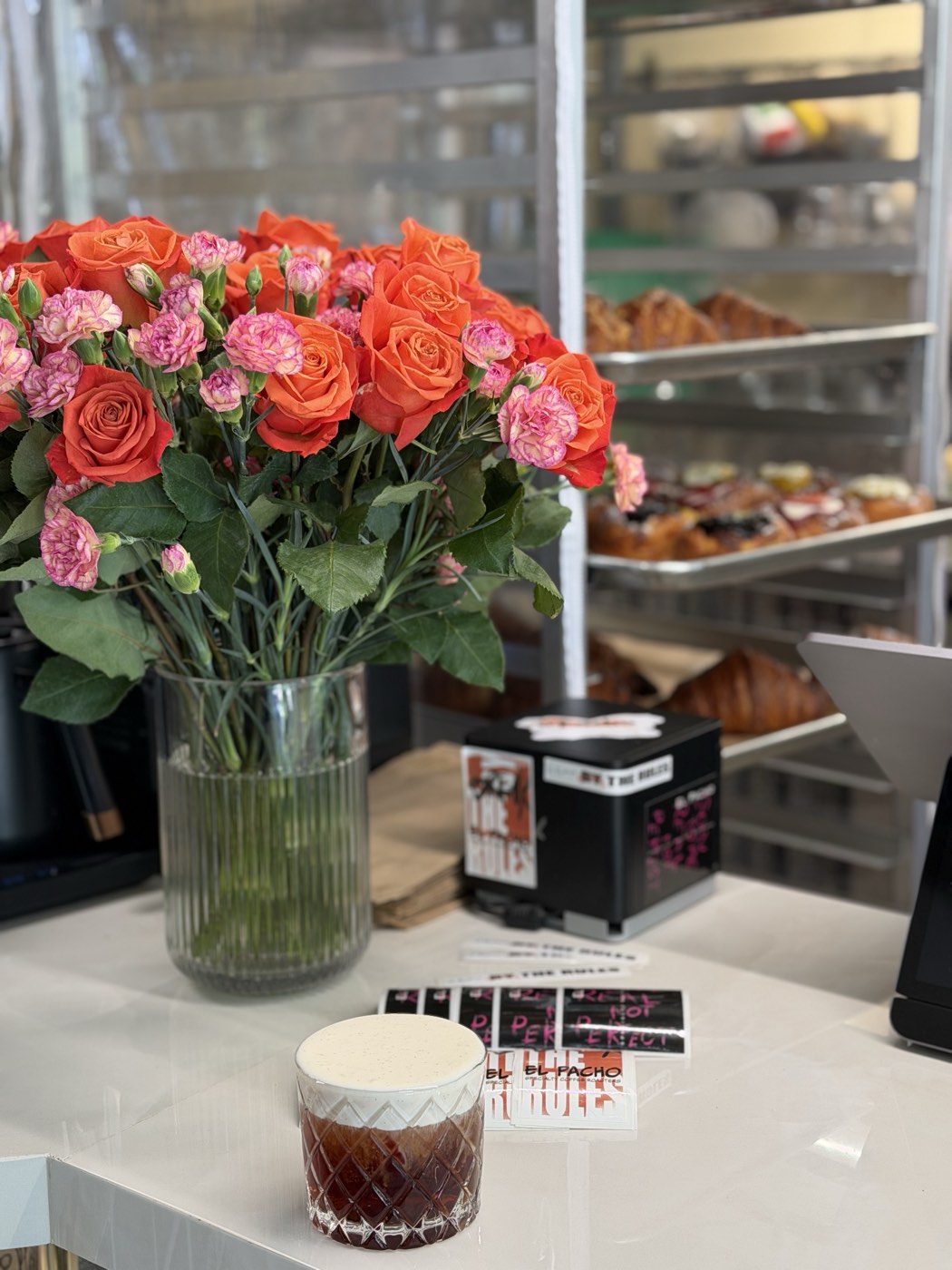 A vase of orange and pink roses and a cold coffee on El Pacho's counter, with trays of croissants and pastries on the rack behind